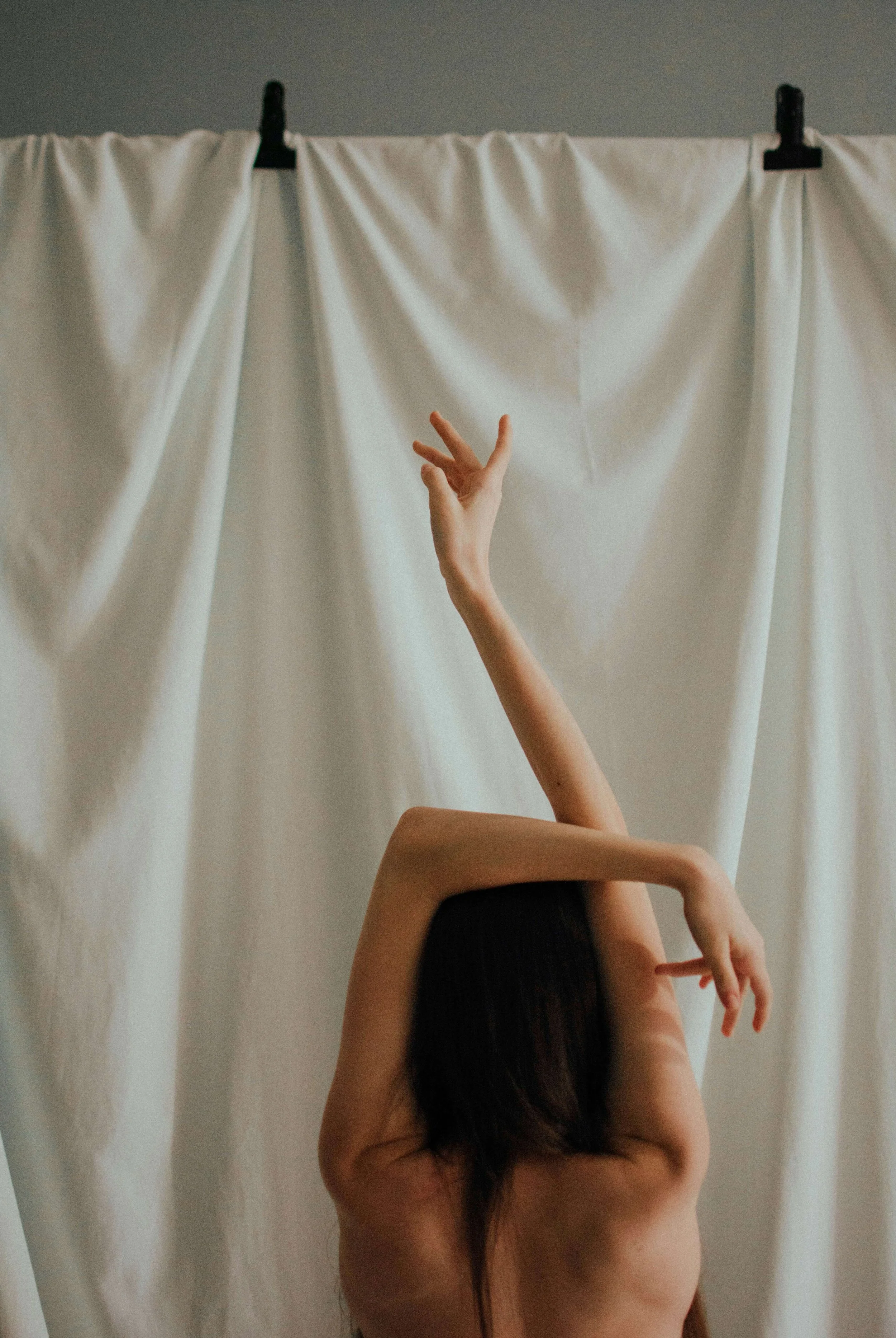 A woman with long dark hair is posing topless against a plain backdrop with white curtains, with one arm bent behind her head and the other arm extended upward with fingers gracefully spread.
