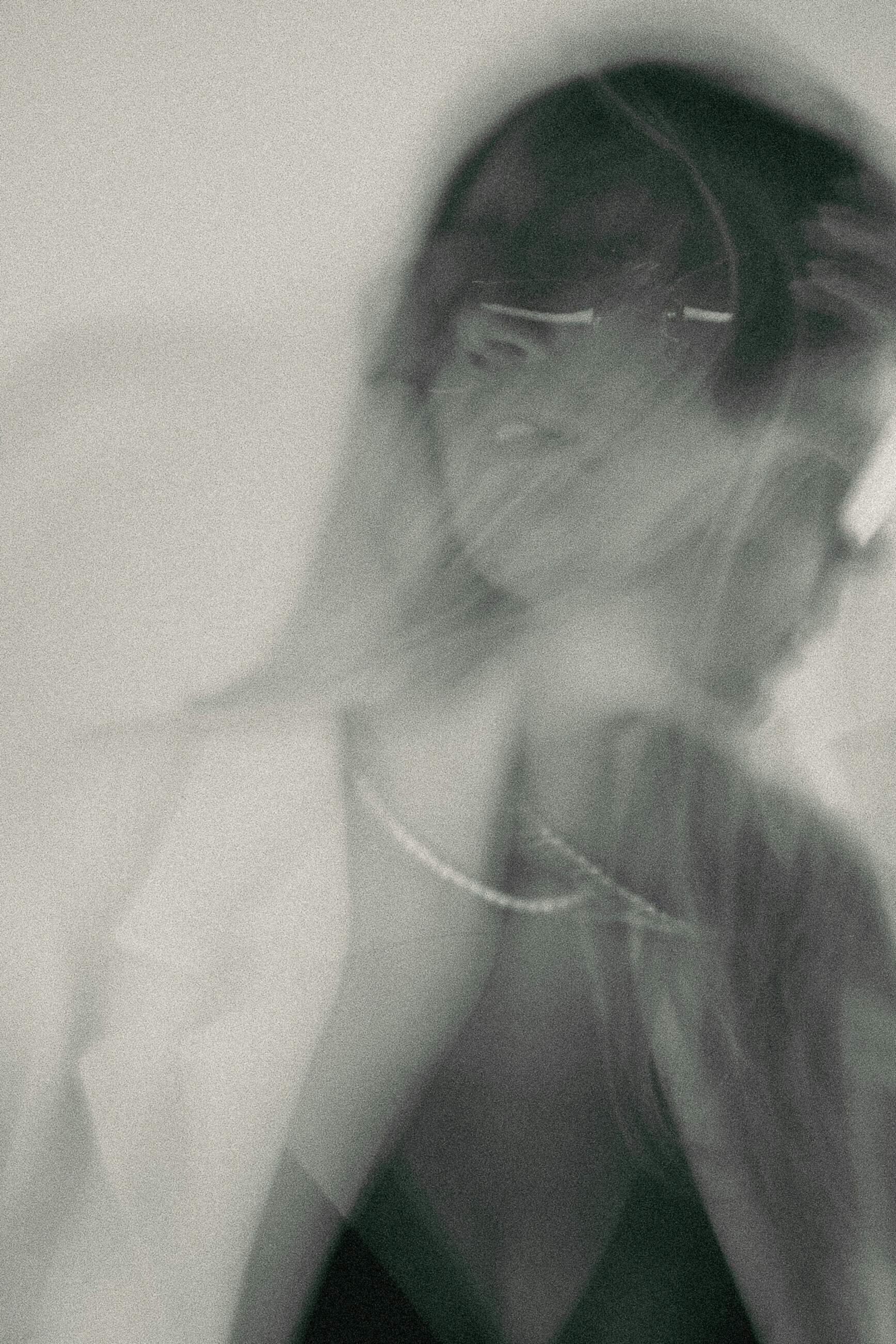 Black and white photo of a woman wearing sunglasses and a pearl necklace, with her hair flowing, against a blurred background.