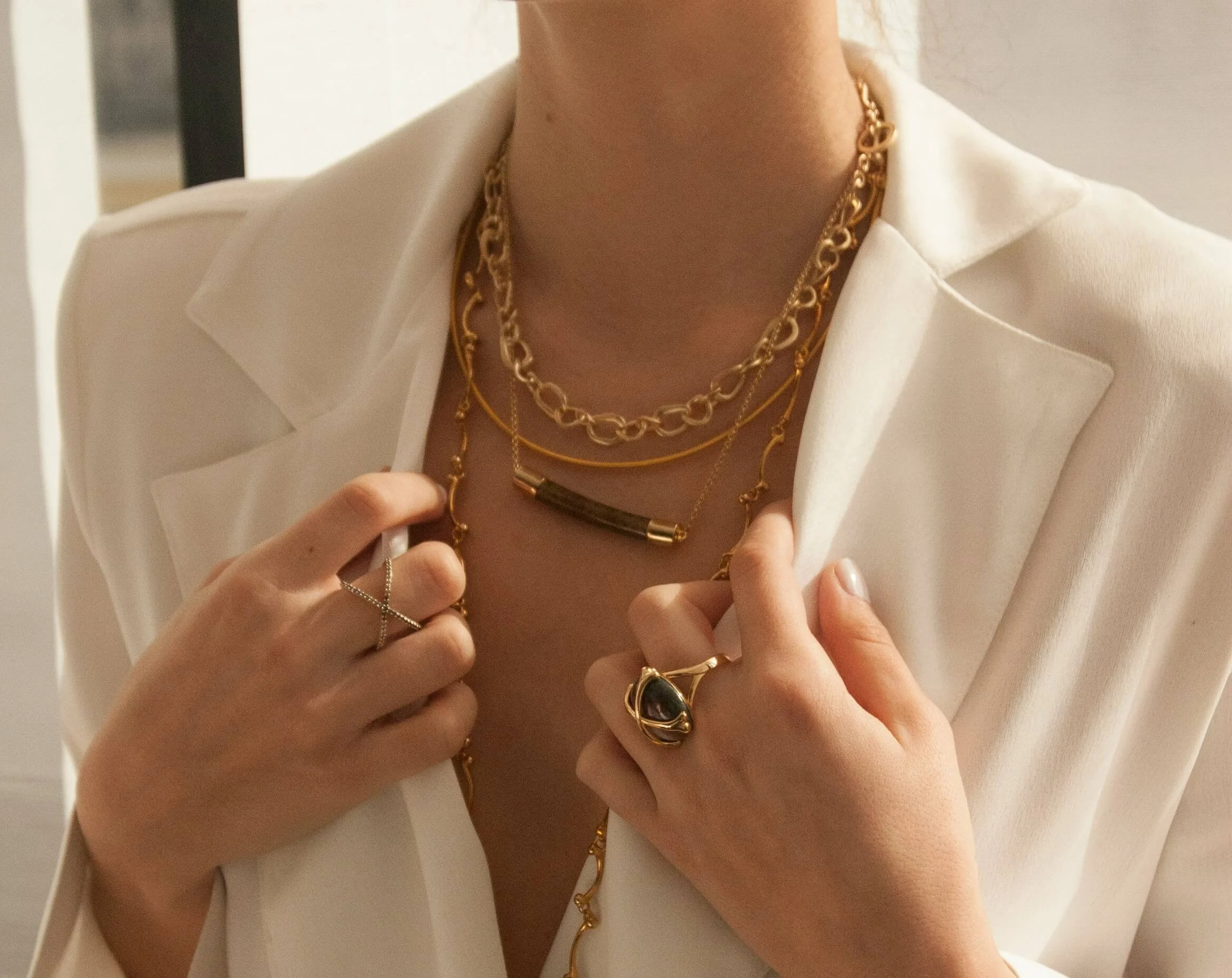 Close-up of a woman wearing layered gold necklaces and rings, dressed in a white blazer.