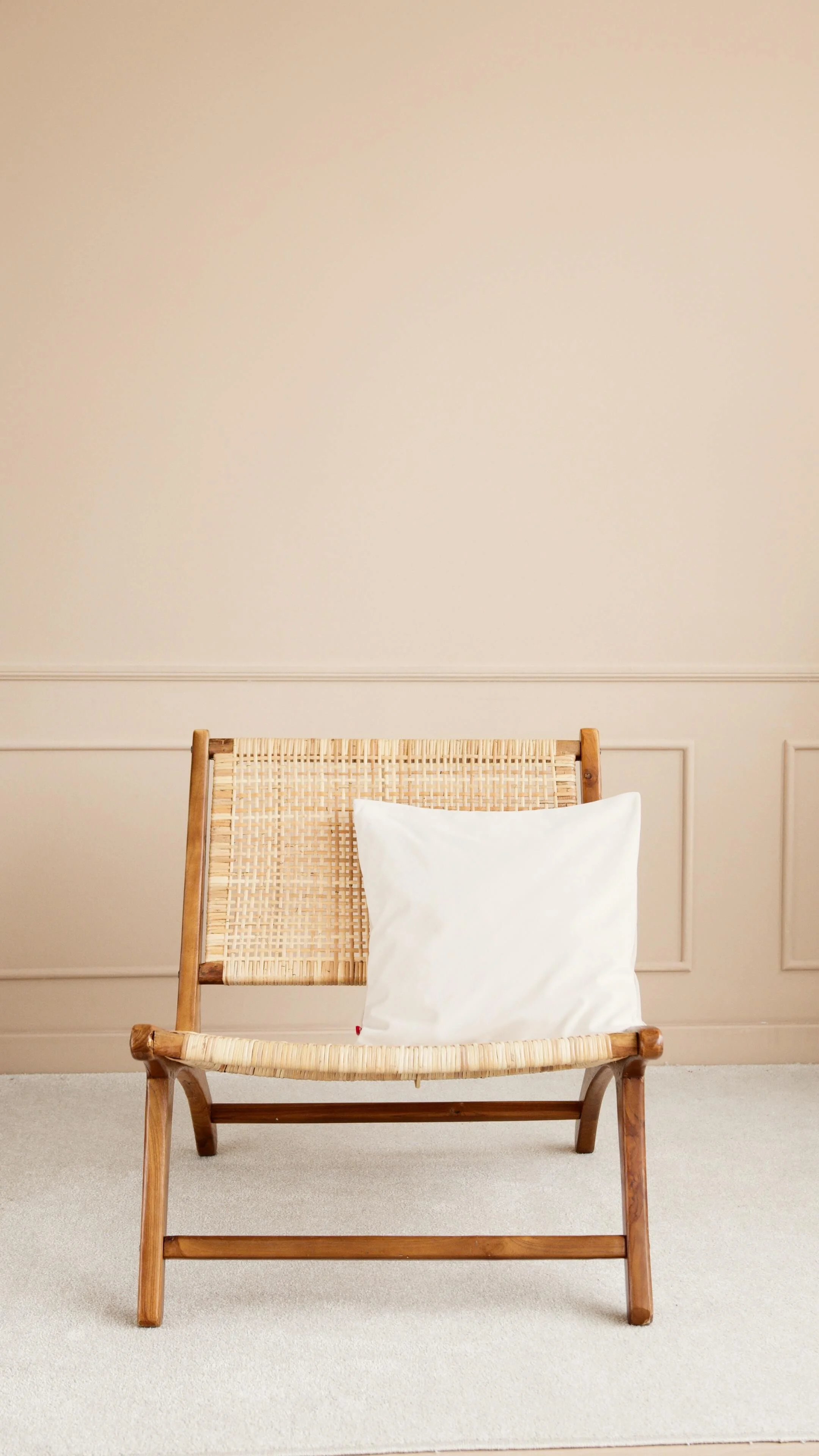 A wooden chair with a woven cane backrest and seat, with a white pillow placed on it, located against a beige wall with decorative paneling.