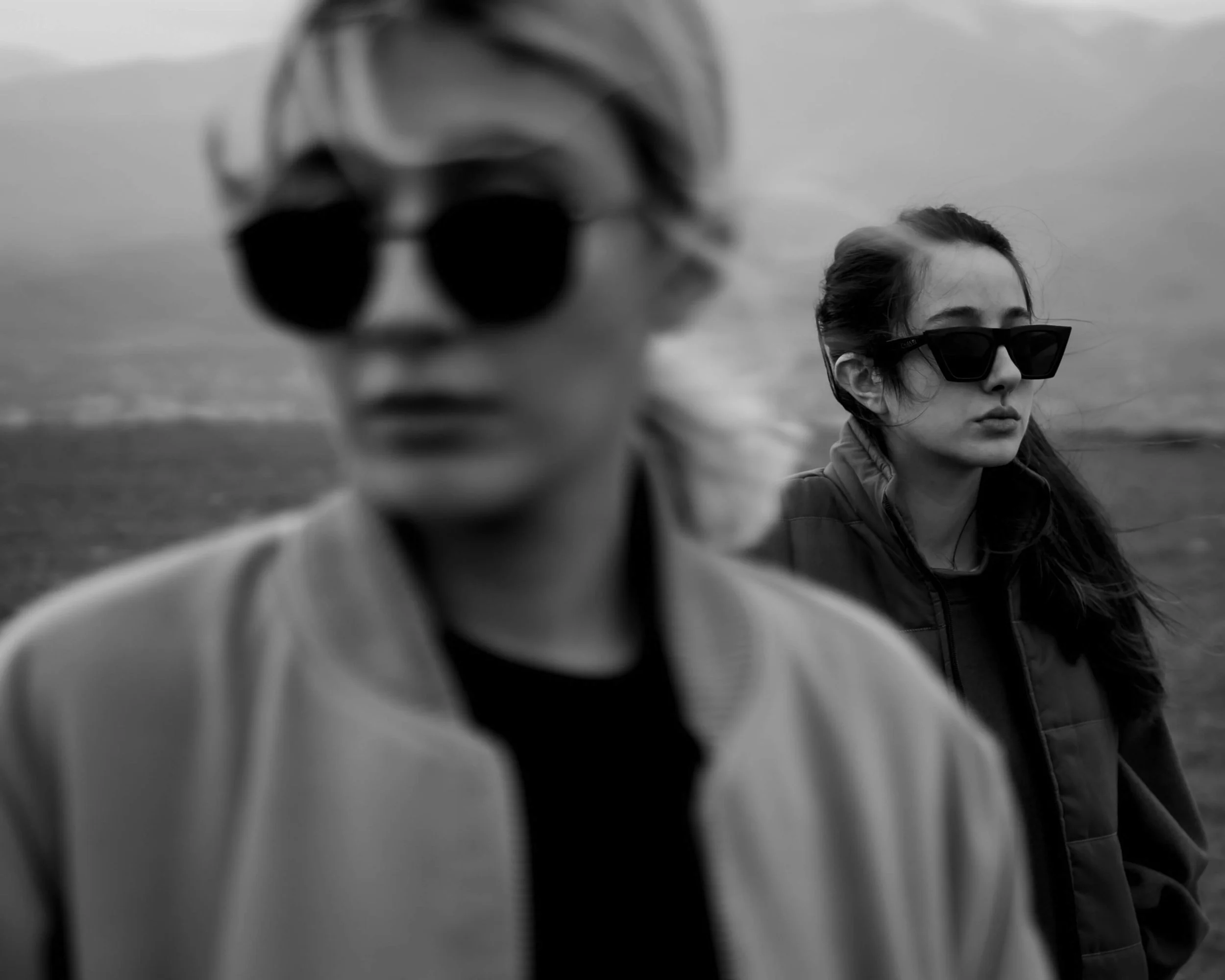 Two women outdoors in black sunglasses, one in the foreground and blurred, and the other in the background with long dark hair, both with serious expressions.