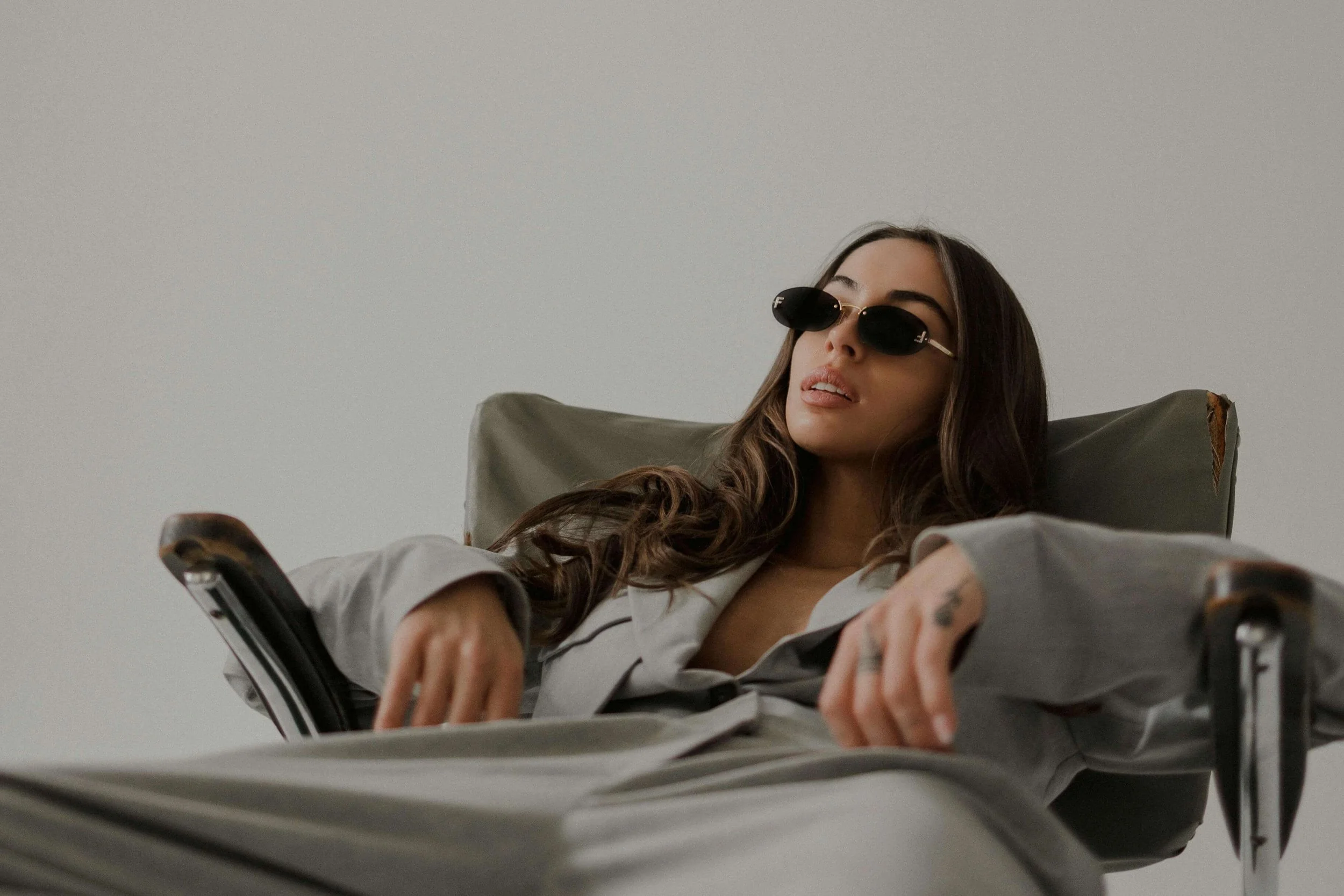 A woman with long brown hair wearing black sunglasses and a gray shirt reclining in a chair with her hands resting on her stomach against a plain gray background.