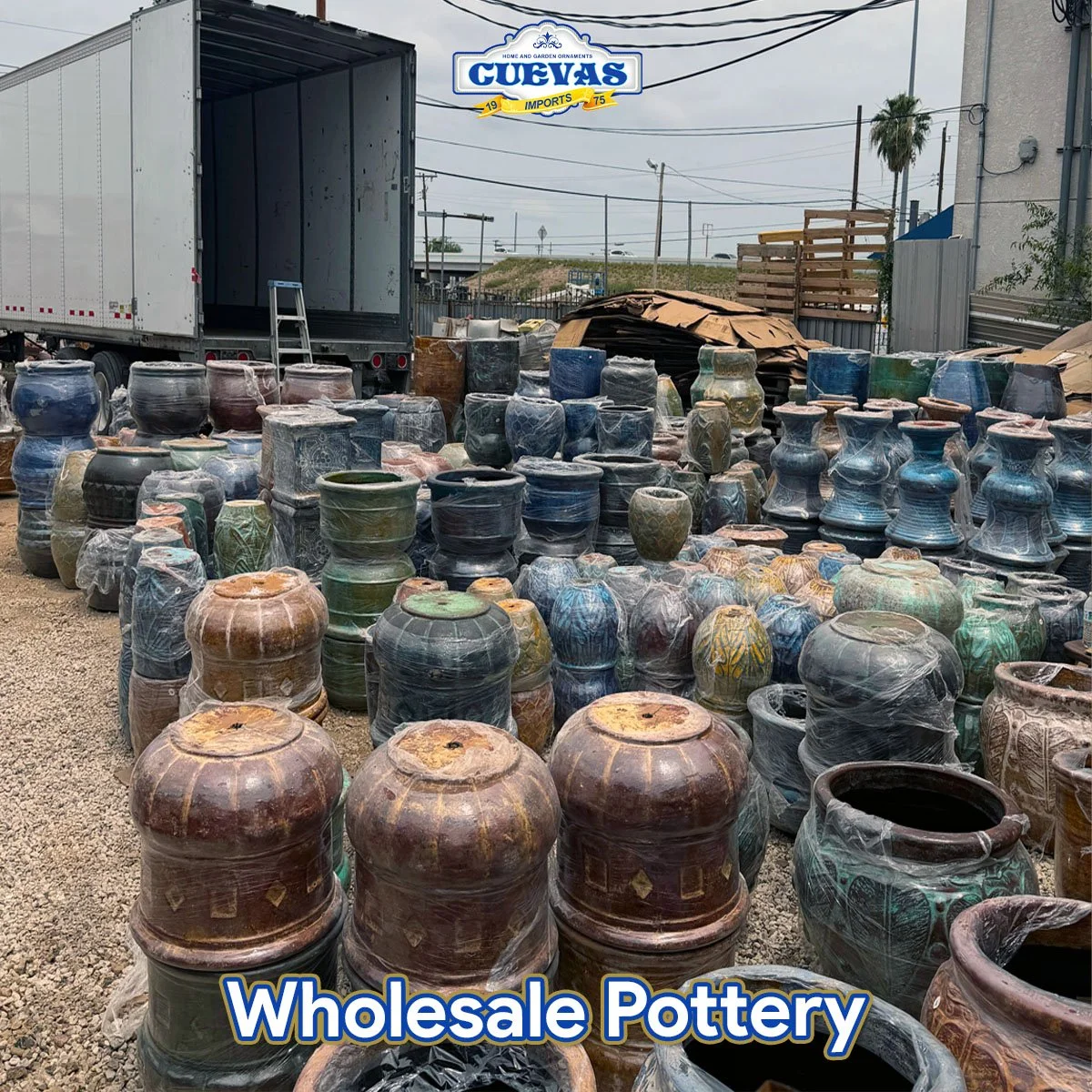 Various colorful pottery and ceramic vases stacked outside at a wholesale pottery store, with a large truck in the background and a sign reading 'CUEVAS Wholesale Pottery'.