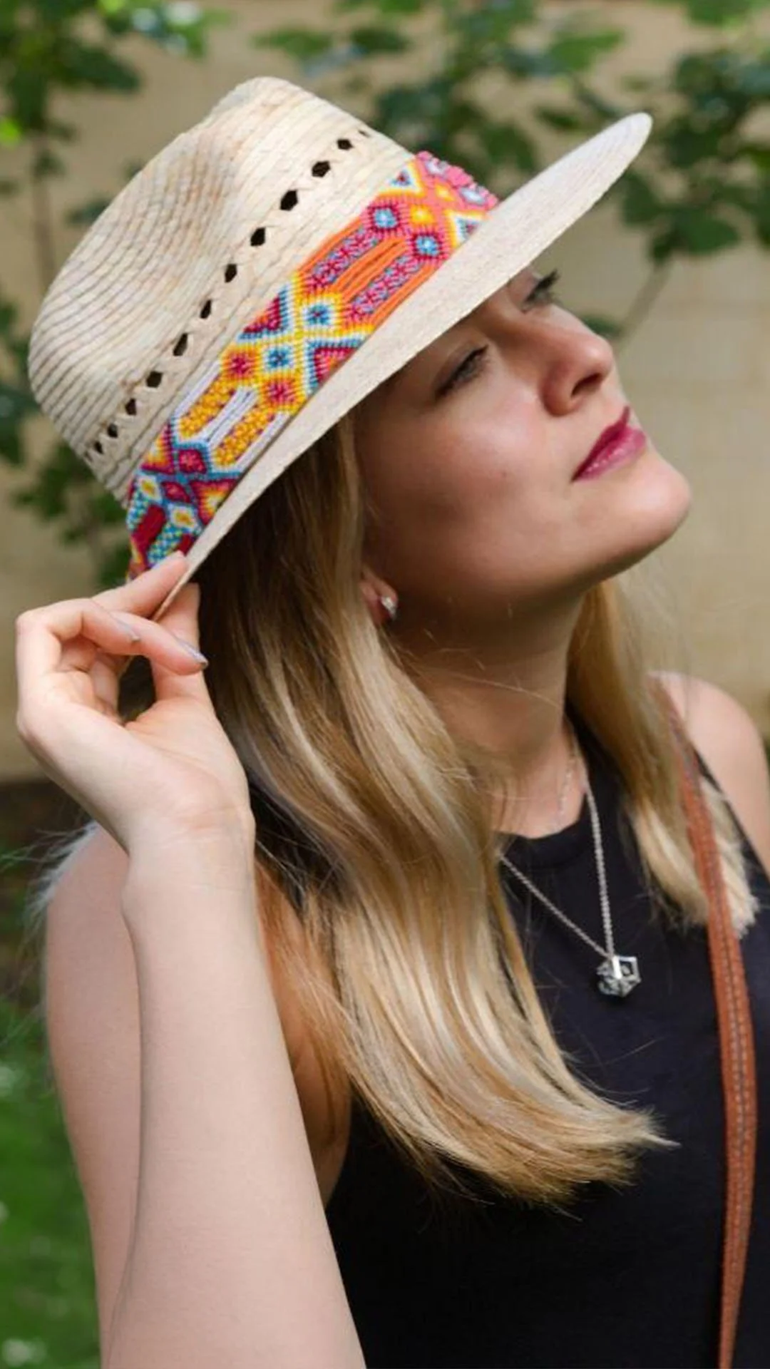 A woman with long blonde hair wearing a wide-brimmed straw hat with colorful embroidery, standing outdoors with green foliage in the background.