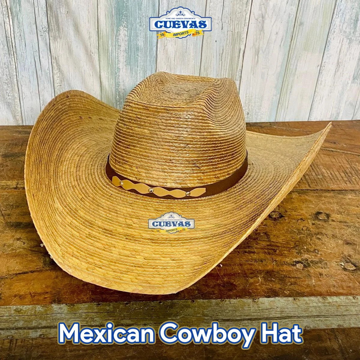 A straw Mexican cowboy hat with a dark brown band around the crown, resting on a wooden surface with a light, weathered wooden wall background.
