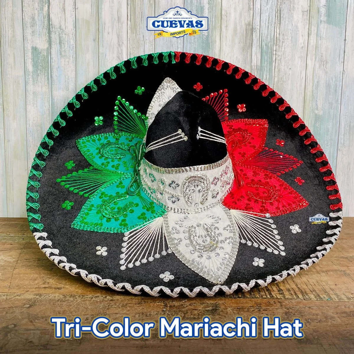 A tri-color Mexican mariachi hat decorated with green, white, and red embroidery, sequins, and designs, displayed on a wooden surface against a light-colored wooden wall.