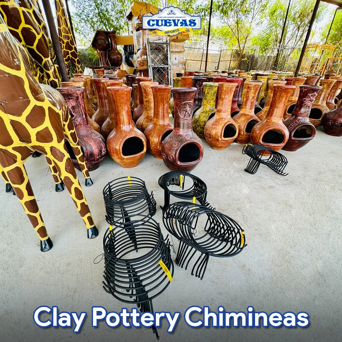 A display of various clay pottery chimneys, some resembling vases with wide openings and others with decorated surfaces, arranged outdoors with a giraffe-shaped sculpture on the left and black metal grill racks in the front. A sign reads "Guevas Imports" in the background.
