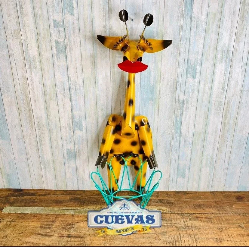 Decorative giraffe made of metal and plastic, standing on a wooden surface against a wooden plank wall, with a sign reading 'GUVAS imports 1975' in front.