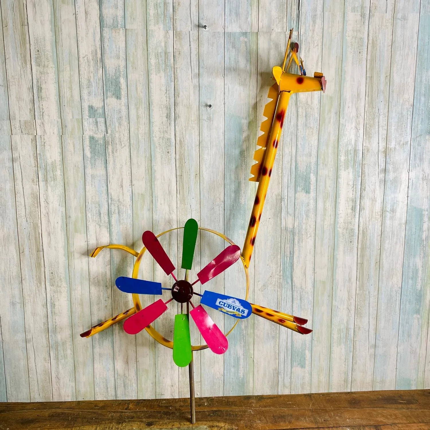Colorful pinwheel with red, blue, pink, green, and yellow blades on a thin metal rod, positioned in front of a light wood-paneled wall.