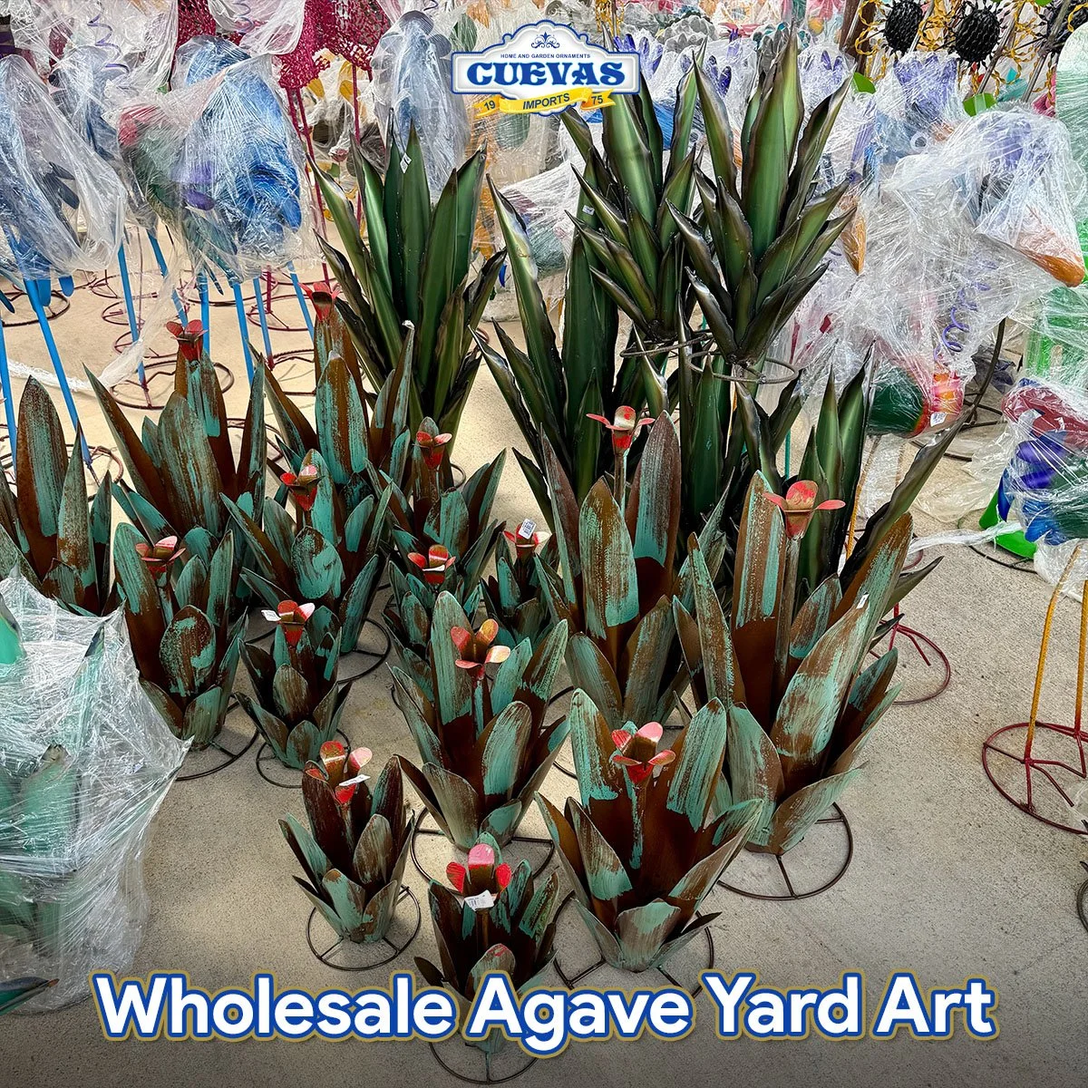 Display of artificial agave plants in various sizes at a wholesale yard, with some wrapped in plastic and others standing on metal stands.