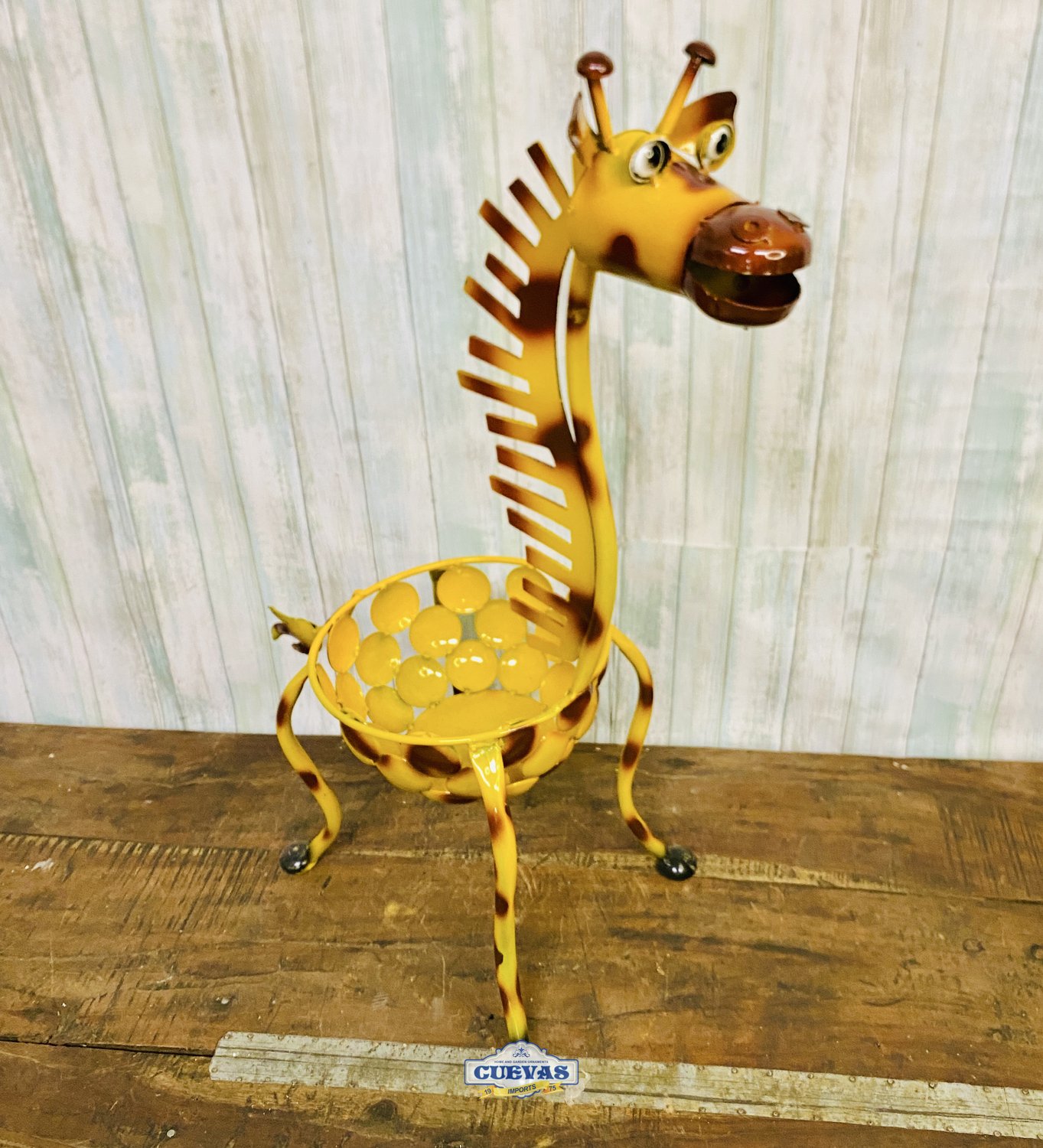 Decorative metal sculpture of a giraffe with a yellow body, black spots, and a shell for a base; the giraffe has a playful, cartoonish face with large eyes and a wide-open mouth.