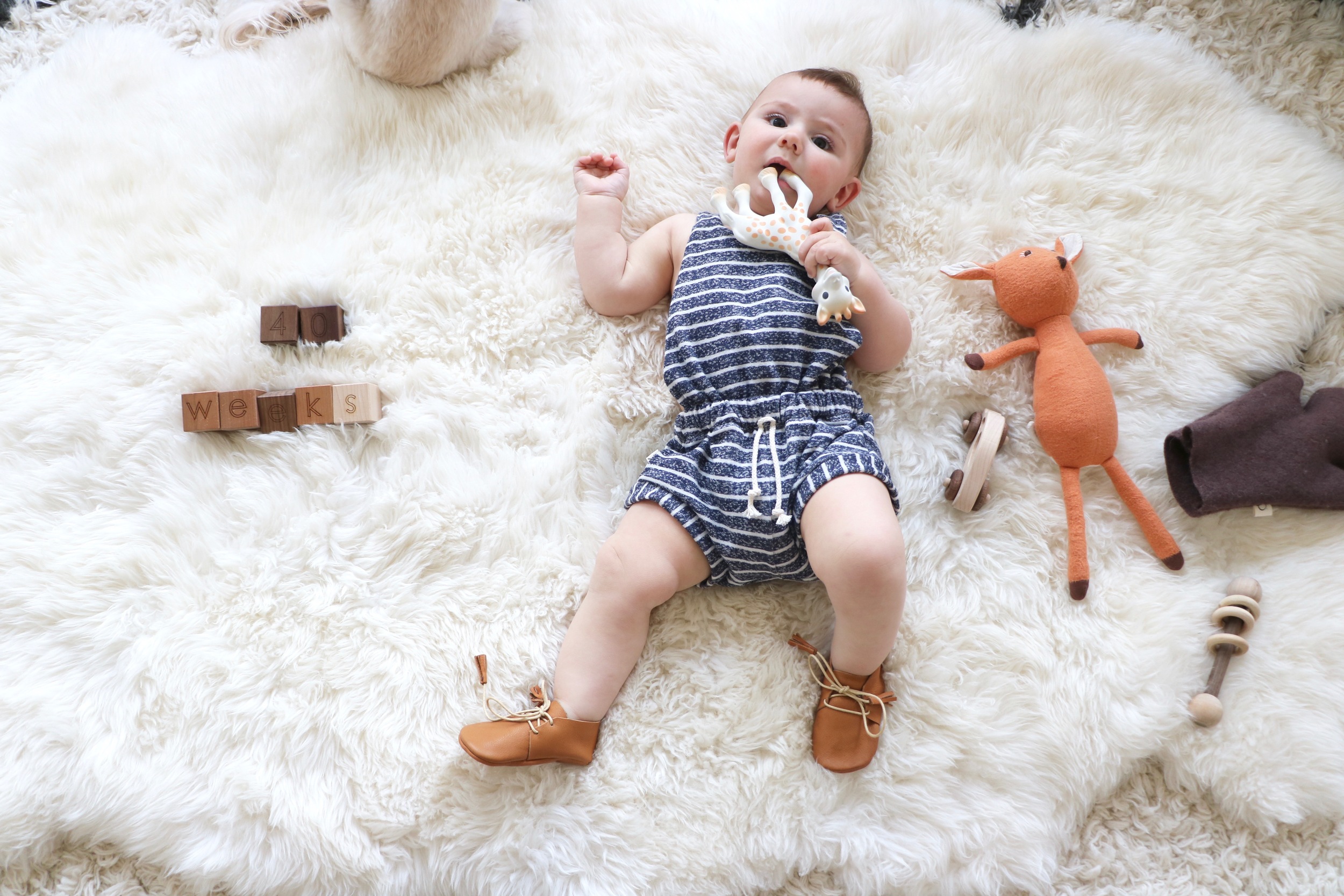 Noam is wearing a Childhoods Clothing romper and Little Pine Outfitter Scout moccasins . Our blocks are from Little Sapling Toys and his fawn doll is Hazel Village .