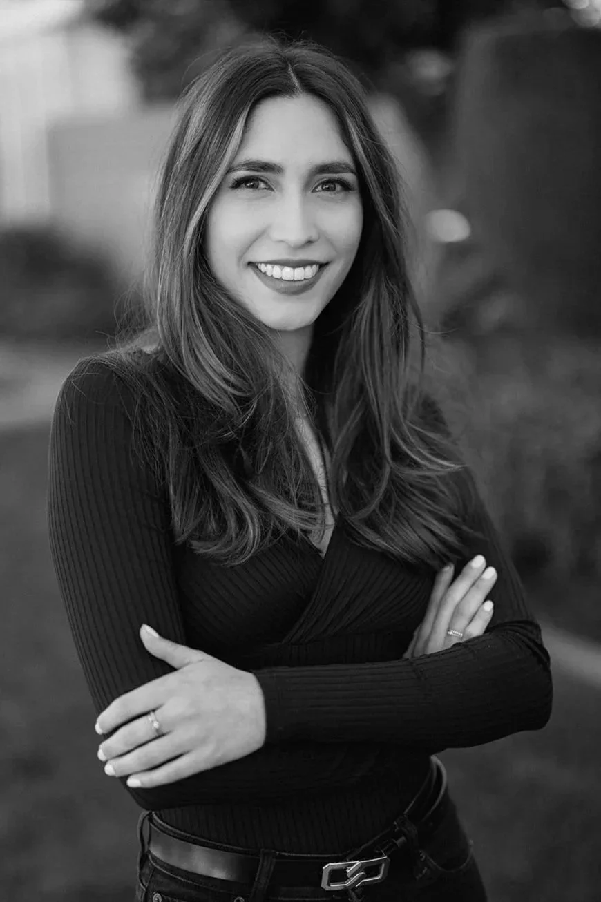 A black and white photo of a young woman with long hair, smiling, wearing a long-sleeved shirt, with arms crossed, outdoors in front of a blurred background.