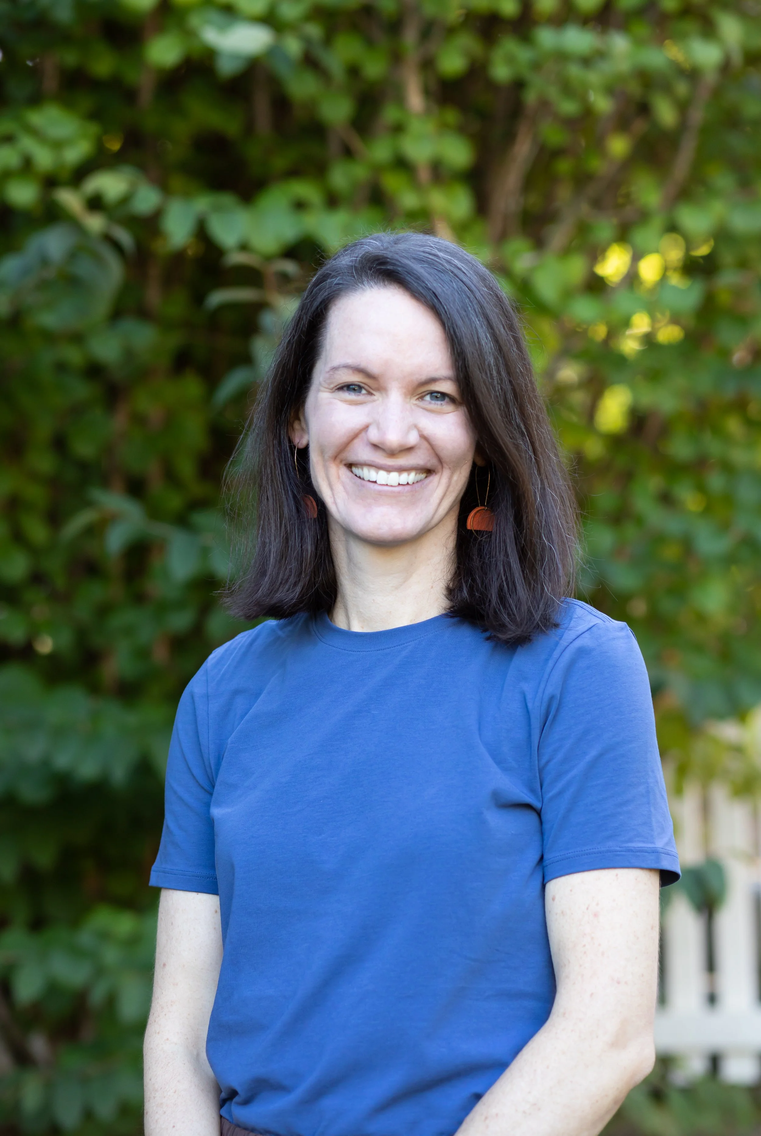 A woman with dark hair and light skin, smiling and wearing a blue shirt, standing outdoors in front of green foliage.