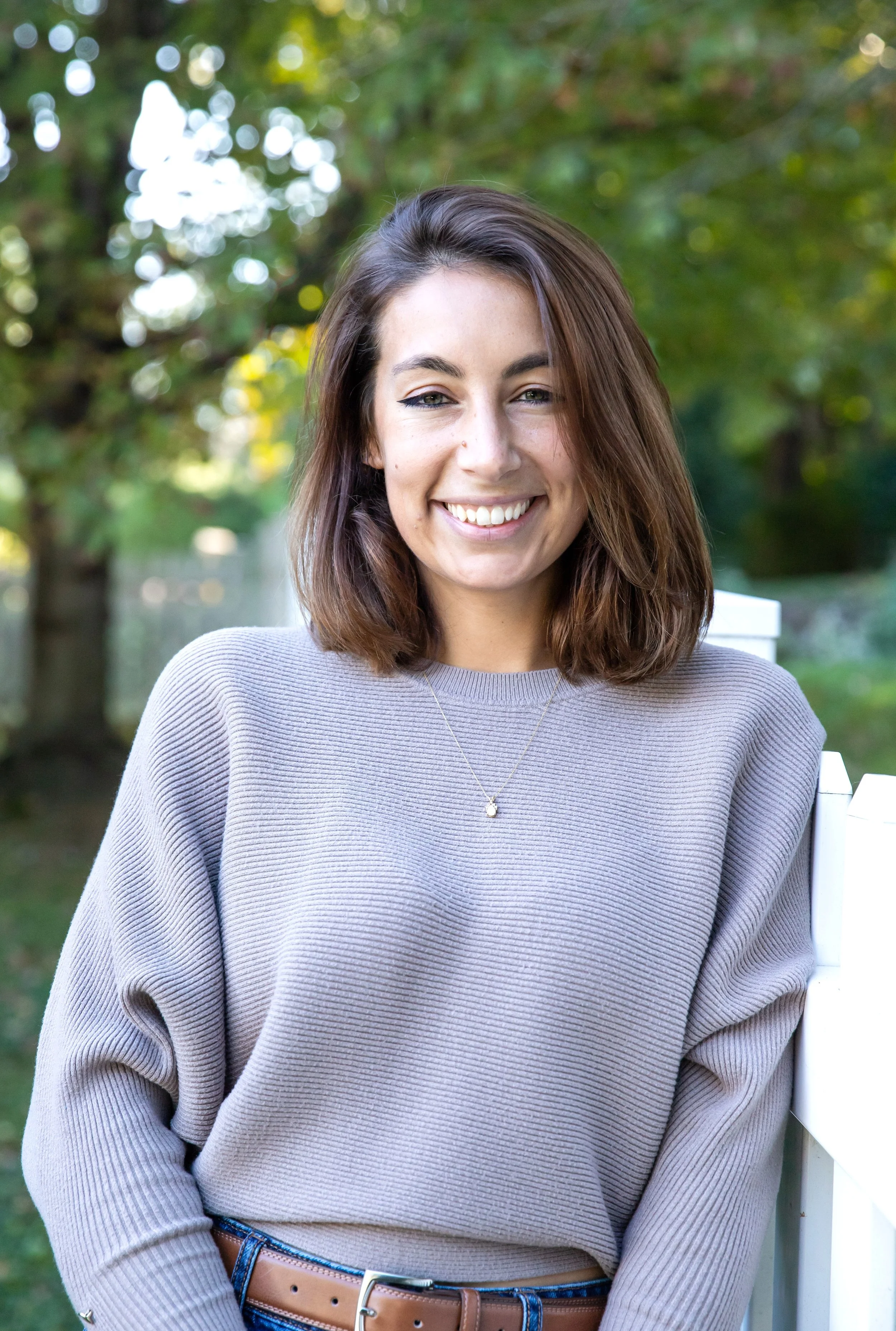 Smiling young woman with brown hair wearing a light gray sweater and necklace, outdoors in a garden with green trees in the background.
