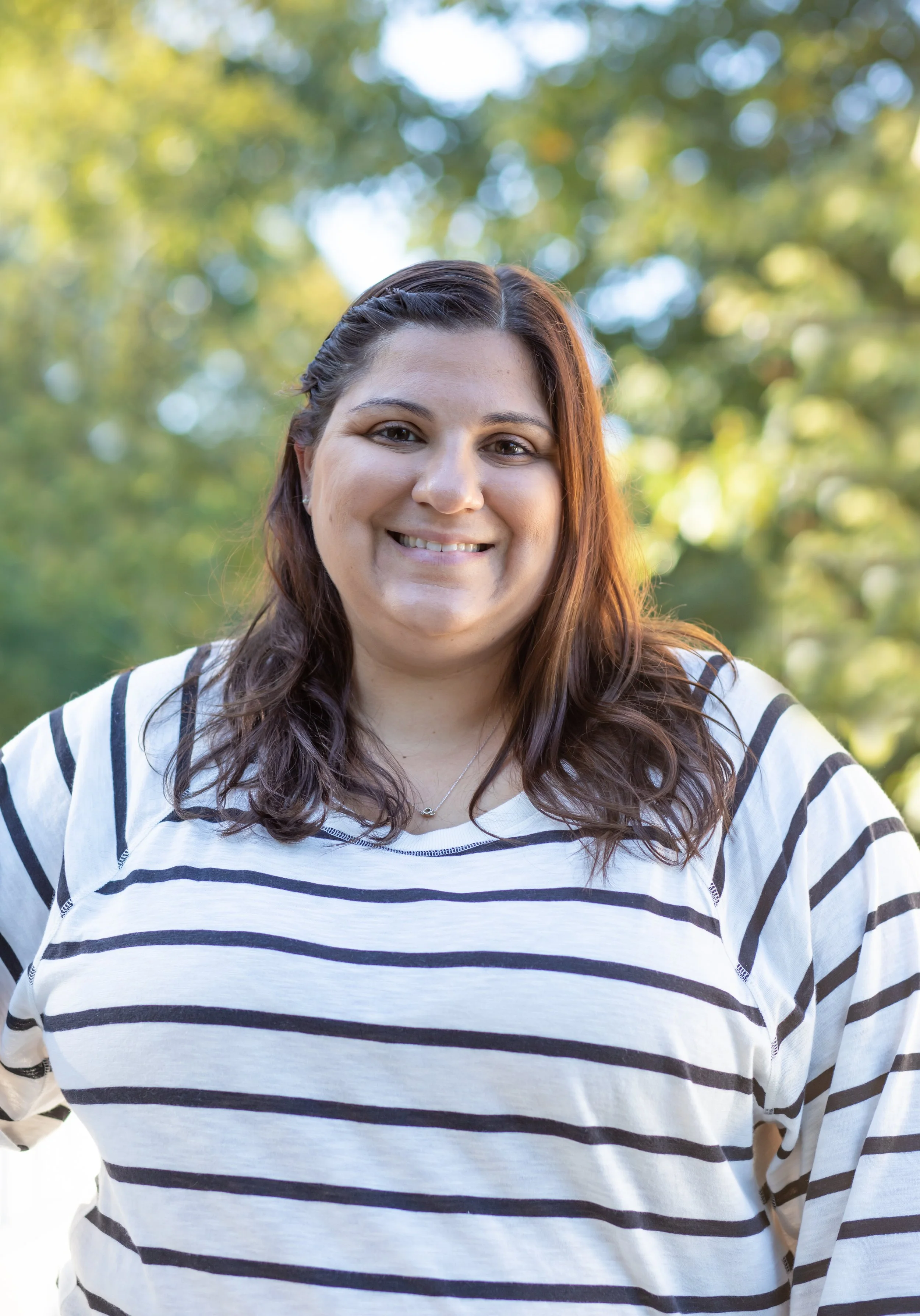 A woman with shoulder-length brown hair and a white and black striped shirt smiling outdoors with blurred trees in the background.