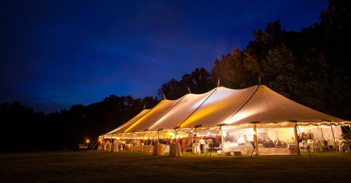 A large white wedding tent in a field during the nighttime. Bright lights illuminate the tent for the guests inside.