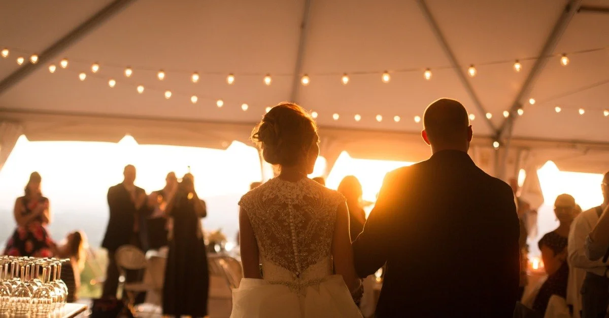 A bride and groom entering a crowded wedding tent on a sunny day. String lights hang above the couple.