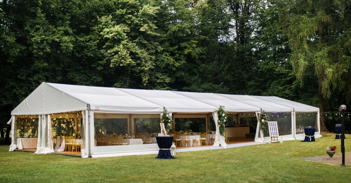A long white wedding tent full of tables and lights set up in front of many massive trees during the daytime.