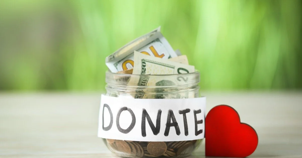 A small jar full of dollars and coins next to a plastic red heart on a table. A sign on the jar says “Donate”.