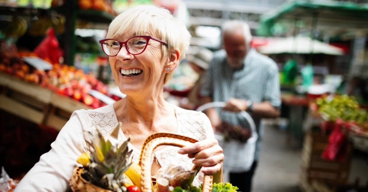 A person wearing glasses smiling with a basket of fruits and vegetables at a farmers market during the daytime.