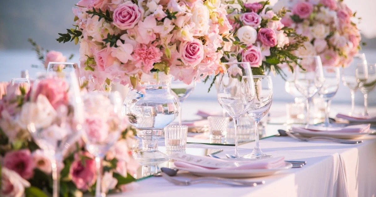 A long white wedding table outside during the daytime. Glassware and many pink flowers decorate the entire table.