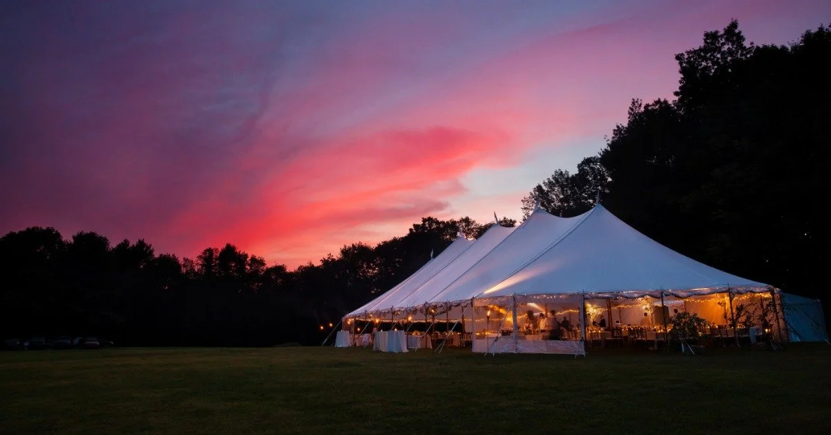 The sun setting over a large white wedding tent outdoors. The tent is brightly lit and full of people.