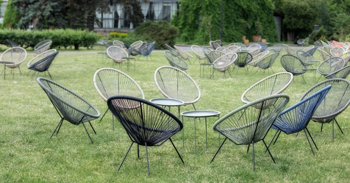 An open field with green grass outside a large home during the daytime. The field is full of black chairs and tables.