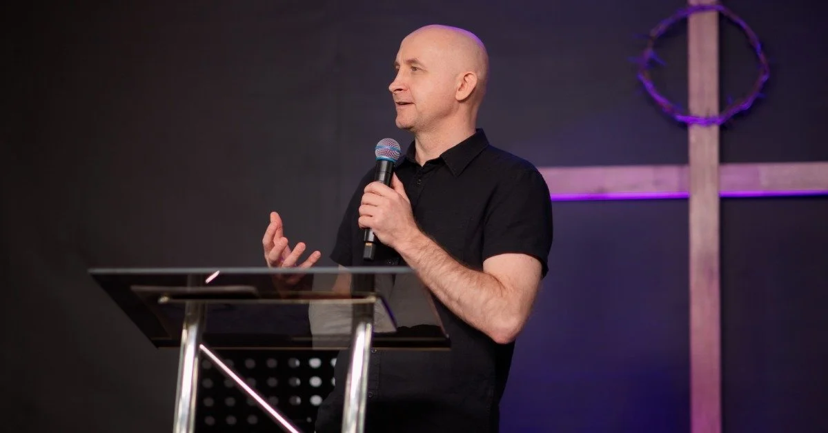 A speaker holding a small microphone in front of a podium on a stage. A cross hangs in the background.