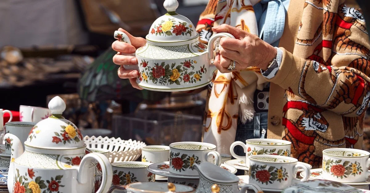 A person holding a porcelain teapot at an outdoor event. The person stands over a table covered in porcelain teacups.
