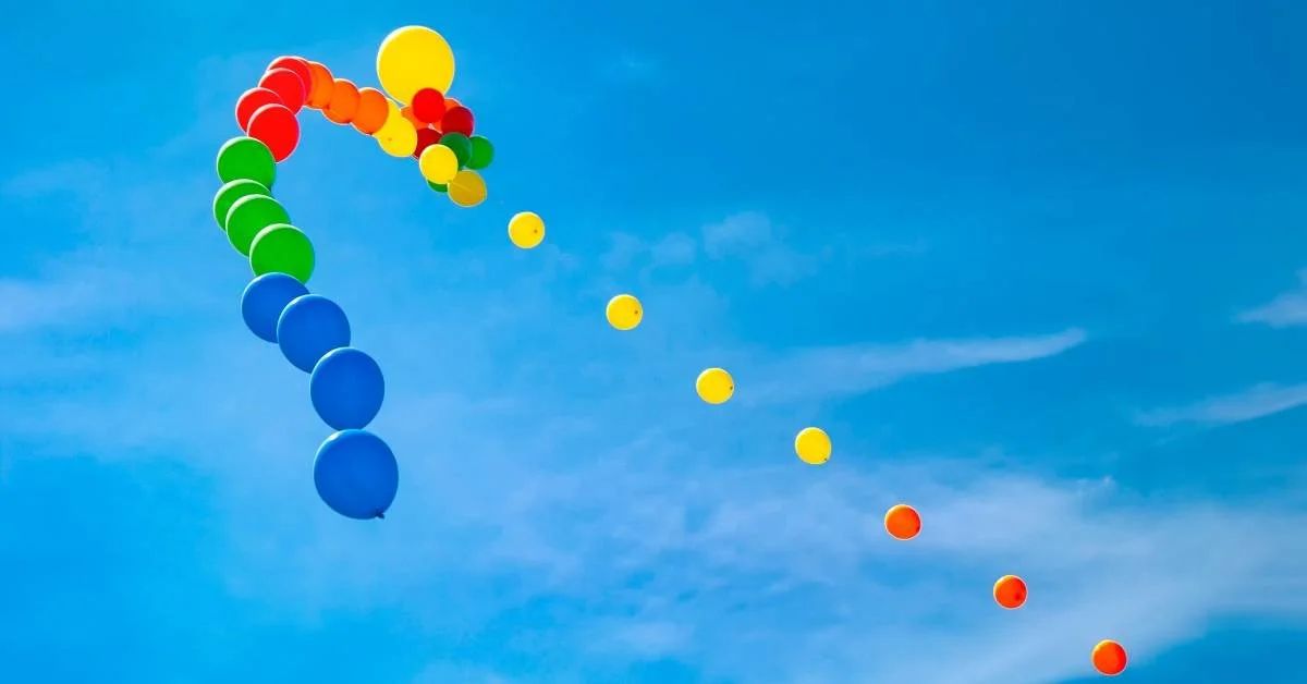 Colorful balloons floating toward the sky on a sunny day. The balloon colors include orange, yellow, green, and blue.