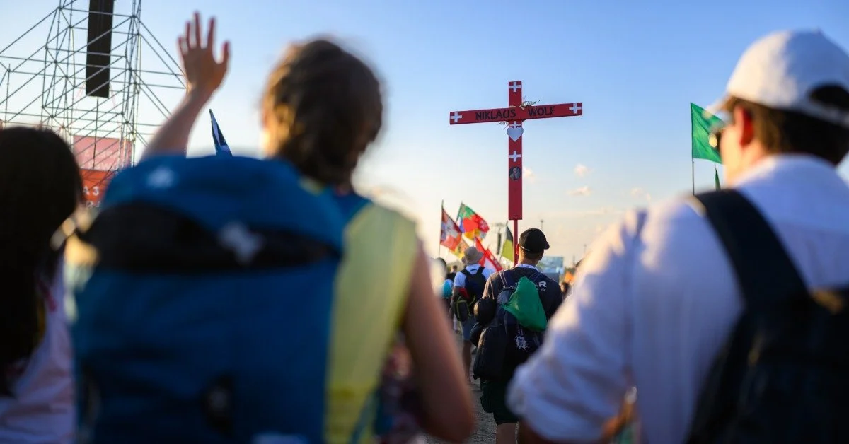 A massive crowd gathering at an outdoor event during the daytime. A large cross is raised in the distance.
