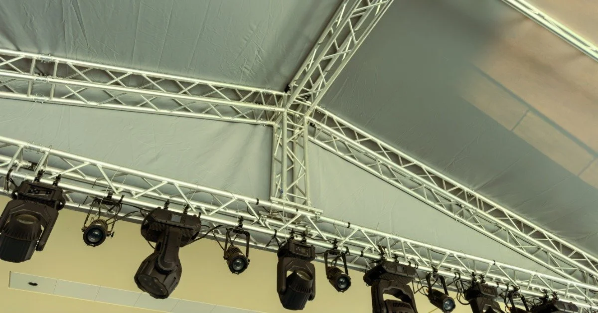 A long row of event lights installed near the ceiling of a tall white tent. All the lights are turned off.
