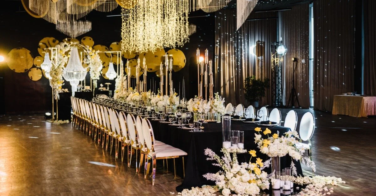 A long banquet table covered in glassware, floral displays, and a black tablecloth. Many chairs surround the table.