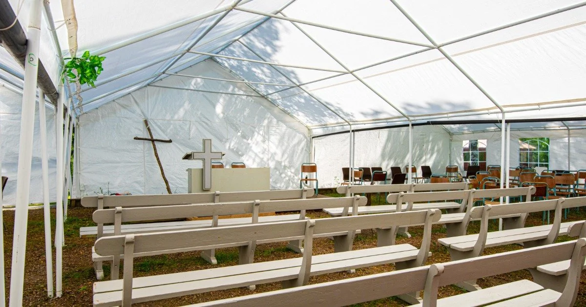 A large white tent on a sunny day. A cross, pulpit, and visitor seating area fill up the interior of the tent.