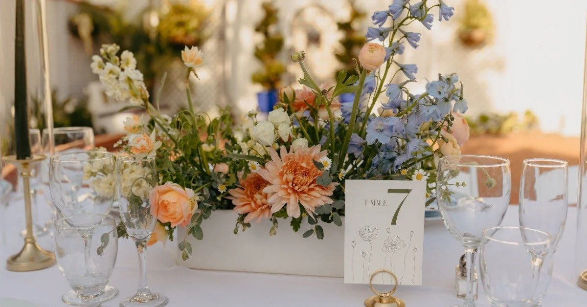 A floral display surrounded by glassware on a table at a wedding. A small sign on the table says “Table 7”.