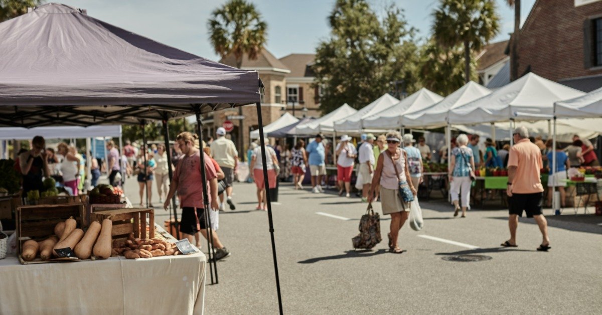 A farmers market full of tents with vendors selling food. One tent is light brown, and the rest are white.