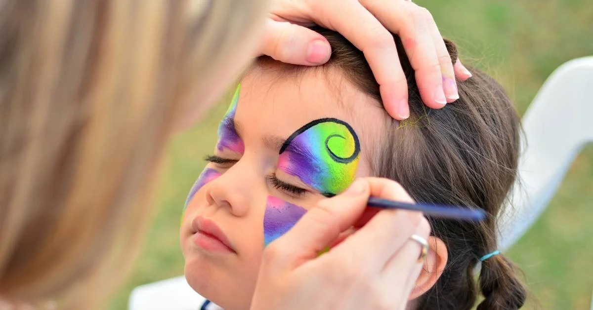 An adult applying face paint around a child’s eyes at an outdoor event. The adult holds a thin paint brush.