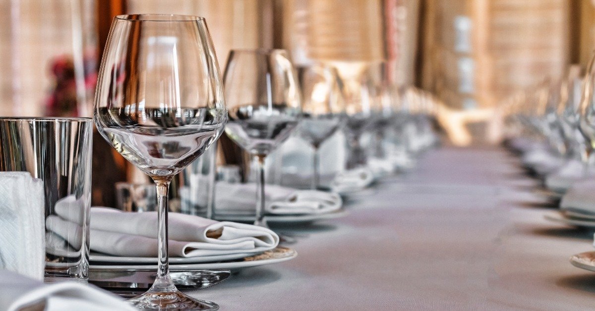 Wine glasses, plates, and silverware neatly organized on a long wedding table in a brightly lit space.