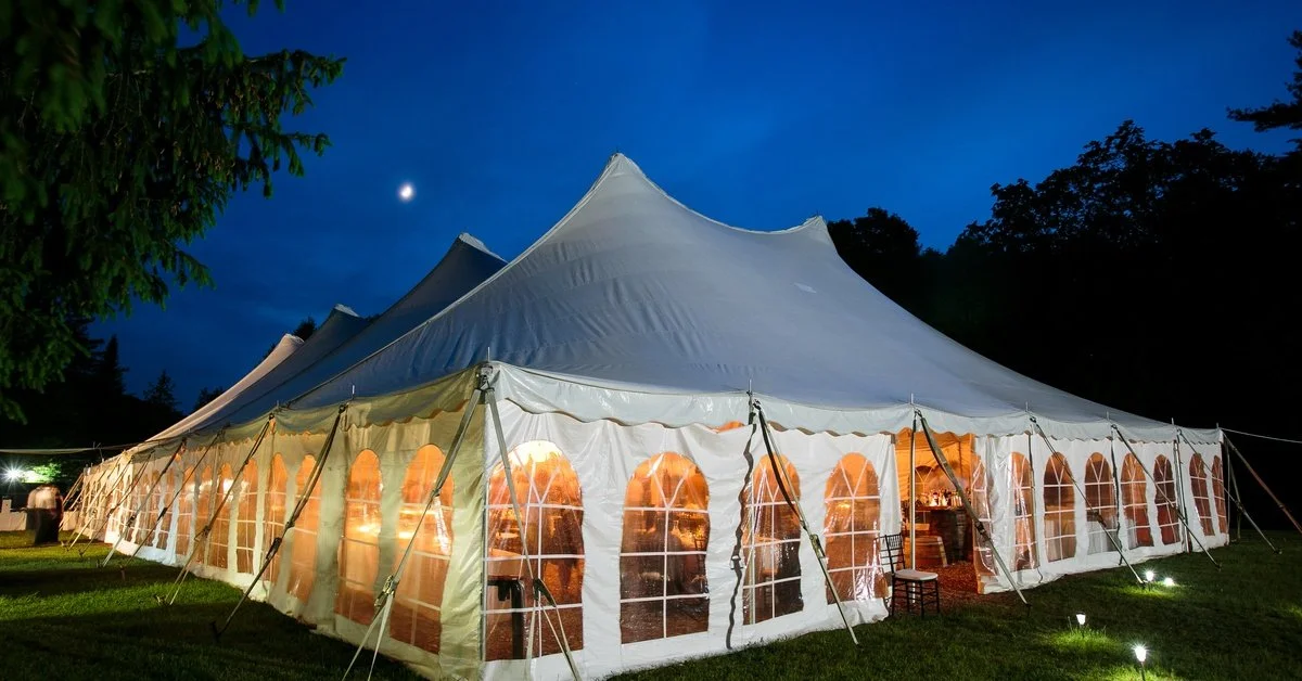 A large white walled event tent with arched windows sits on a field at night. Warm light emits from the tent.