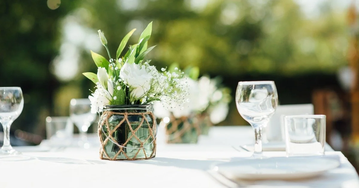 A table outside covered in glassware and a white tablecloth. A row of small glasses with flowers line the table.