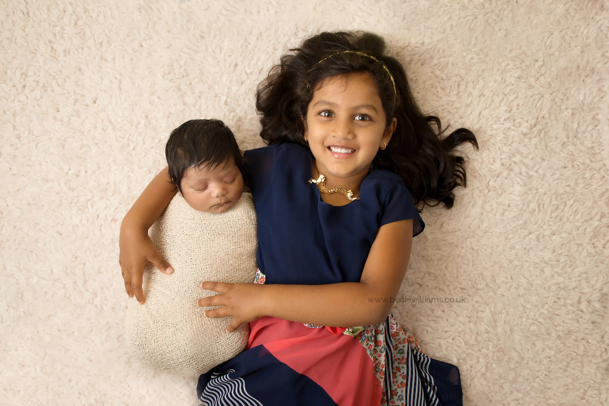newborn baby laying in her big sisters arms on a cream blanket at their photo shoot in hertfordshire