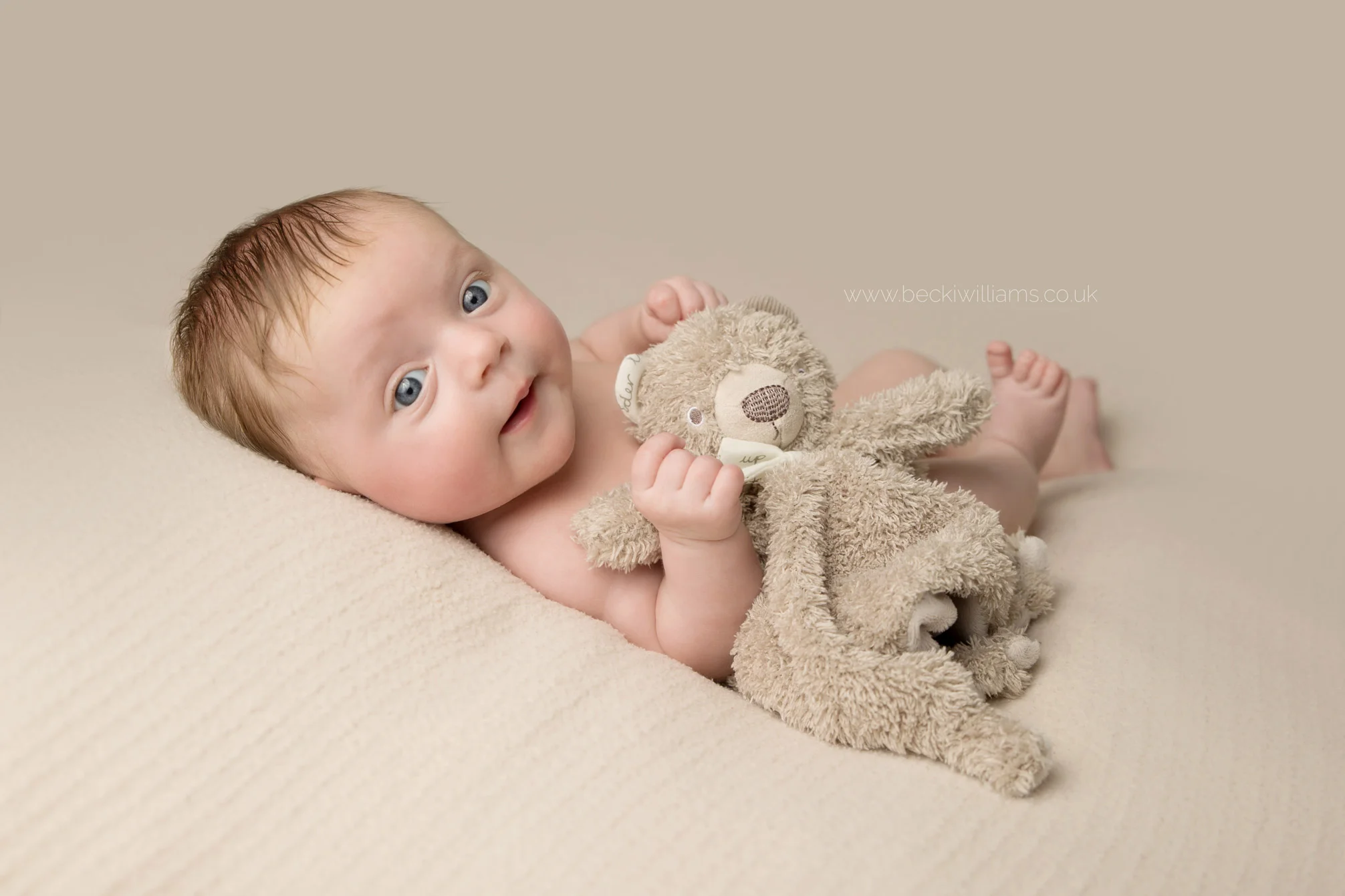 7 week old baby girl lays awake on a cream blanket staring into the camera. she is hugging her brown toy bear comforter and looks happy.