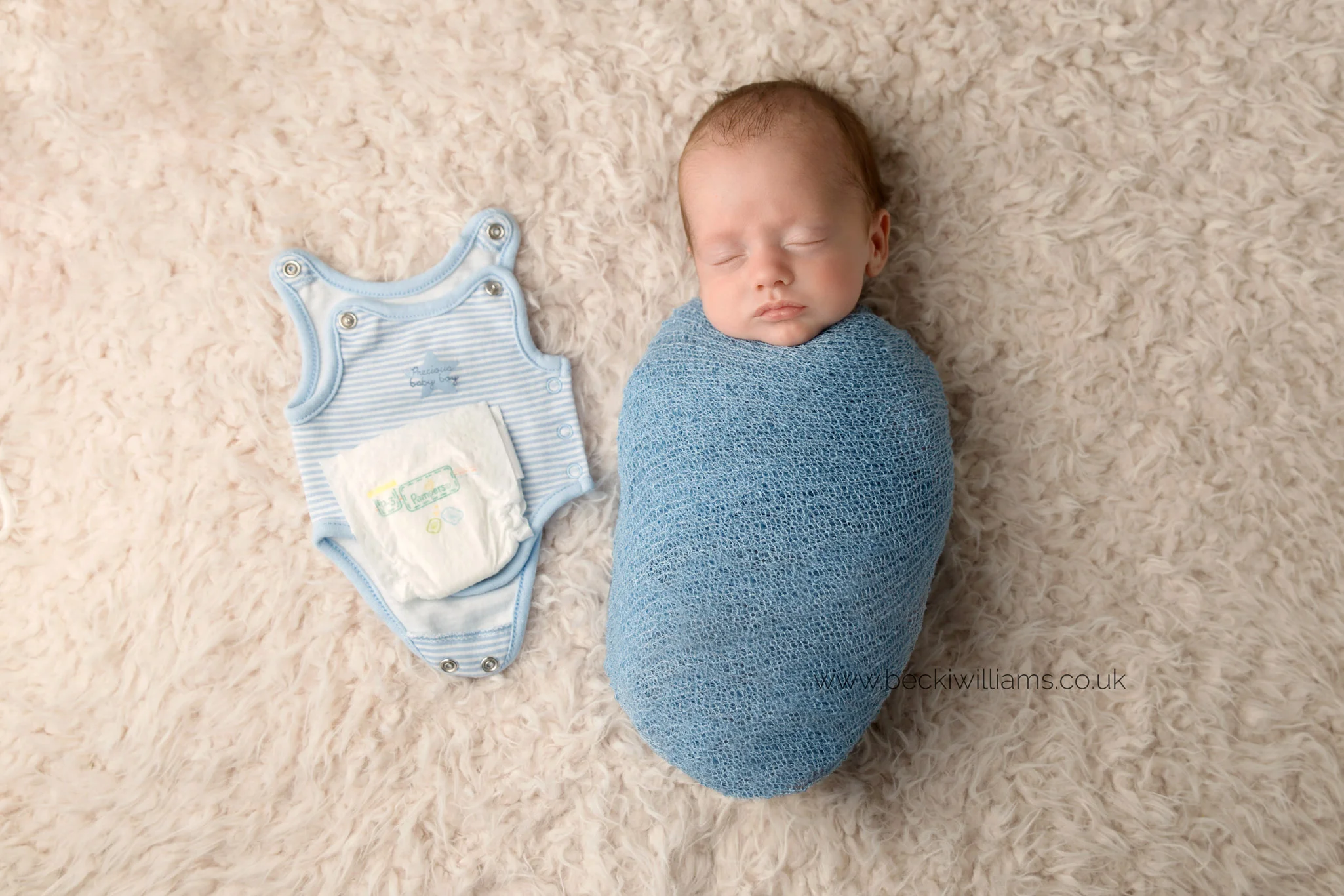 newborn boy lays wrapped in a blue wrap on a cream fluffy blanket. next to him is a tiny baby grow and preemie nappy