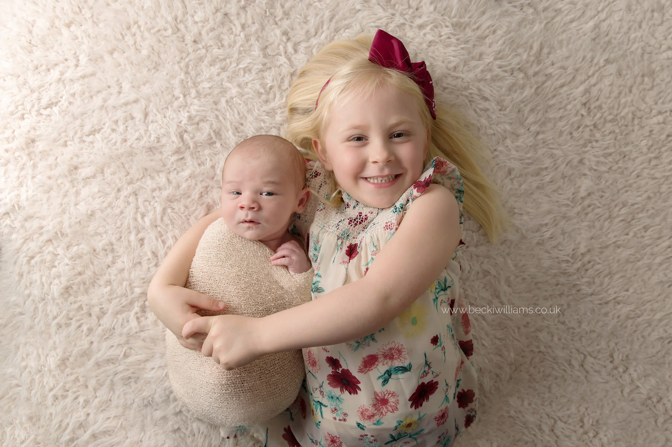 smiling big sister cuddles her newborn baby brother on a cream fluffy blanket