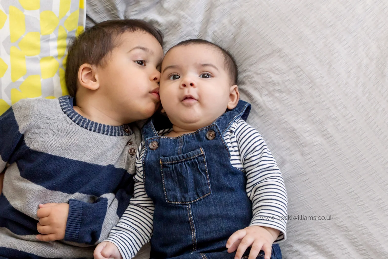 2 year old brother kisses his baby sister on the cheek while laying on his parents bed
