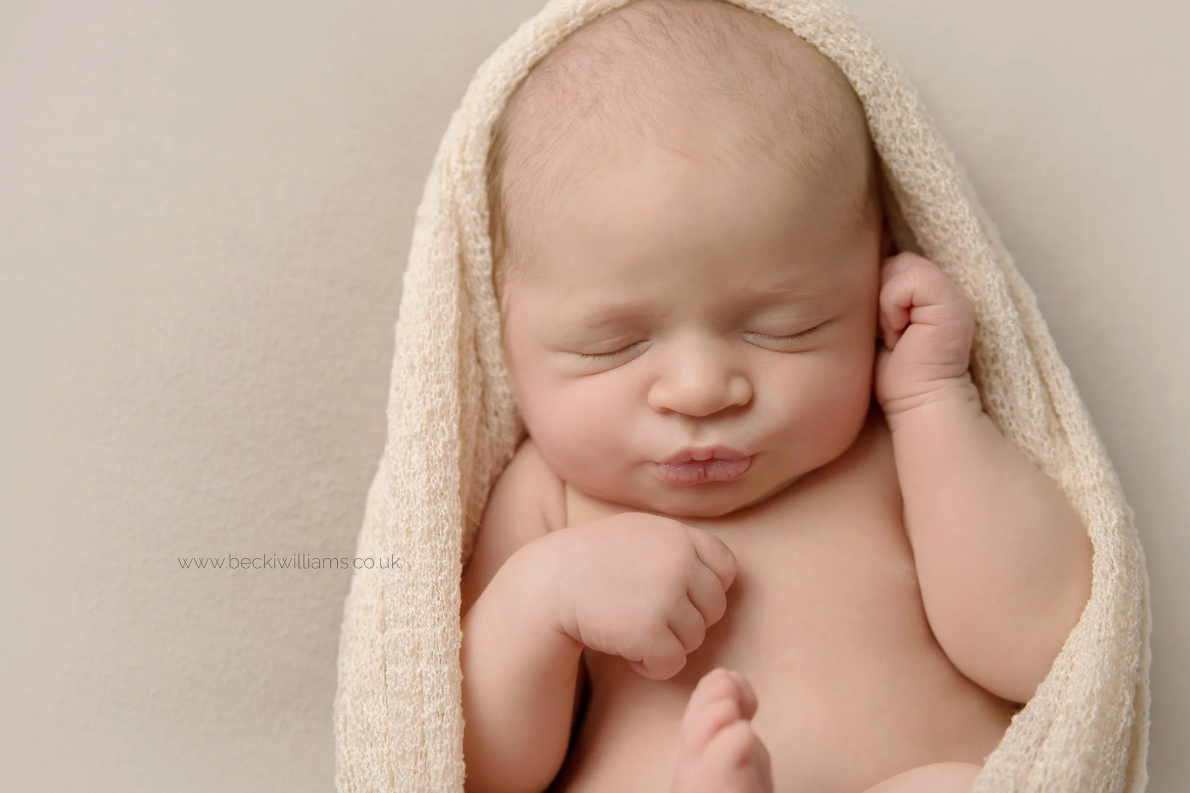 newborn girl laying on her back, pouting, wrapped in a cream blanket