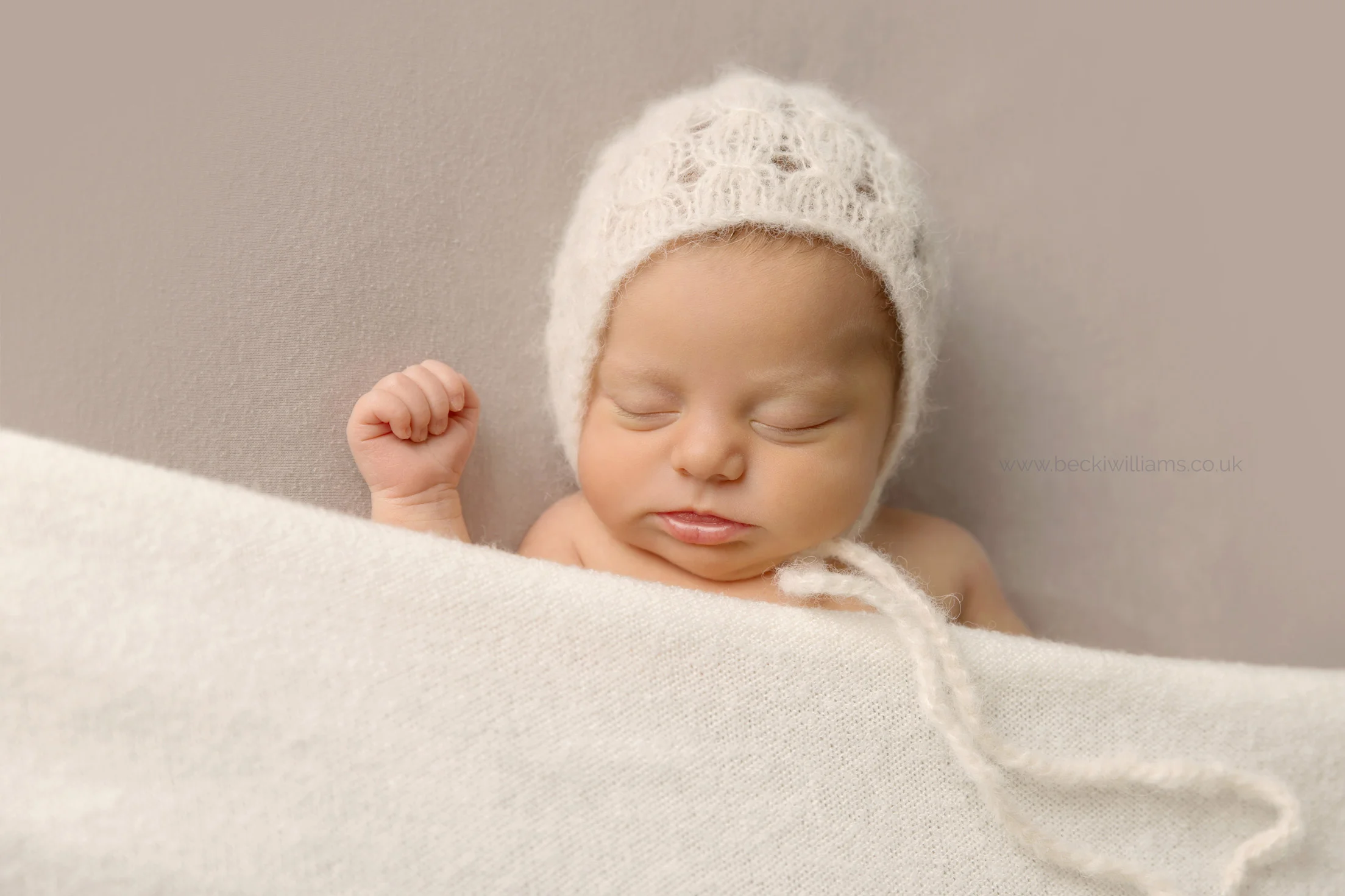 newborn girl asleep under a cream blanket wearing a cream bonnet