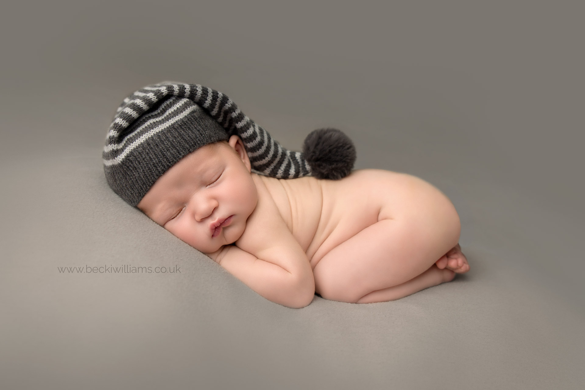 Newborn boy laying sleeping on his tummy on a dark grey blanket, wearing a stripy grey hat