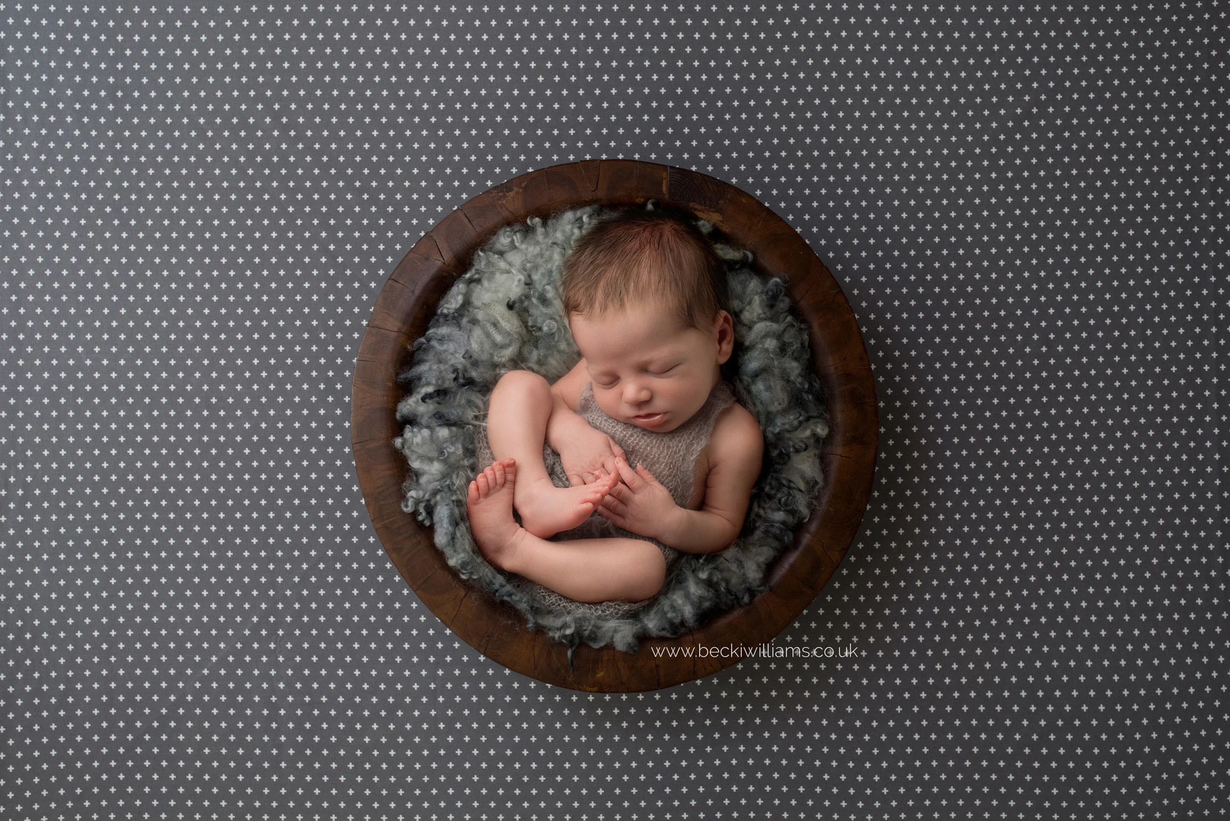 newborn boy lays asleep on grey fur, inside a wooden bowl, which is sitting on a spotty background for his newborn photo shoot in hemel hempstead